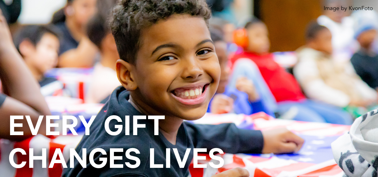 Boy smiles at camera during civics class in Henry Street afterschool program. Text reads, "Every gift changes lives." Image by KvonFoto.